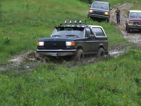 1990 Ford Bronco - A dark green 1990 Ford Bronco navigates a muddy trail, with other vehicles visible in the background.
