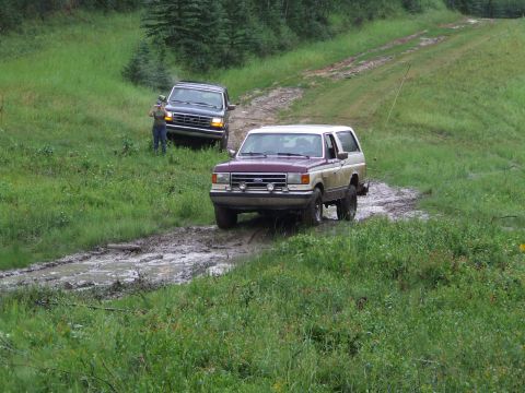 1990 Ford Bronco - Two trucks navigate a wet, muddy path through a grassy, wooded area.
