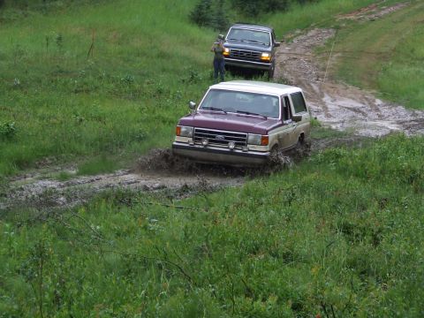 1990 Ford Bronco - Two Ford trucks navigate a muddy trail in a wooded area.