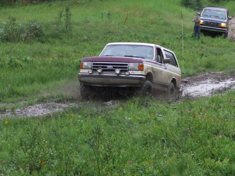 1990 Ford Bronco - A two-tone Ford Bronco navigates a muddy track, with another SUV following in the background.