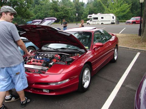 1995 Ford Probe - A red coupe with its hood open is parked in a lot, with a man examining the engine. Other vehicles and an RV are visible in the background.