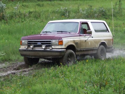 1990 Ford Bronco - A maroon and white Ford Bronco navigates a muddy, grassy terrain.