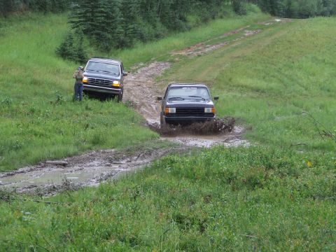 1990 Ford Bronco - Two trucks navigate a muddy trail in a rural, wooded area during daylight.