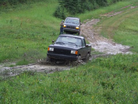 1990 Ford Bronco - Two trucks navigate a muddy off-road trail in a wooded area.