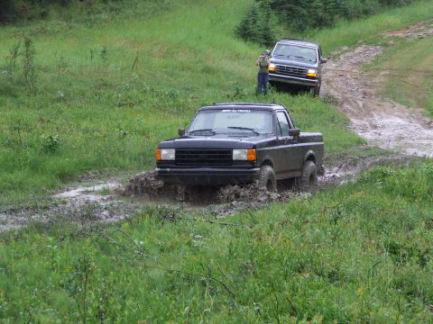 1990 Ford Bronco - Two trucks navigate a muddy trail in a wooded area.