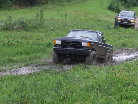 1990 Ford Bronco - Two trucks navigate a muddy track in a grassy, wooded area.