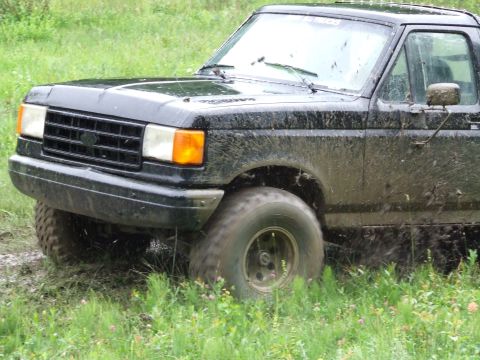 1990 Ford Bronco - Mud-covered black pickup truck navigates grassy terrain.
