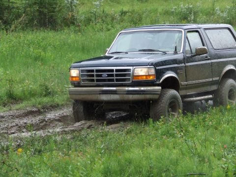 1990 Ford Bronco - A dark blue 1990 Ford Bronco navigates a muddy path surrounded by lush green grass and trees.