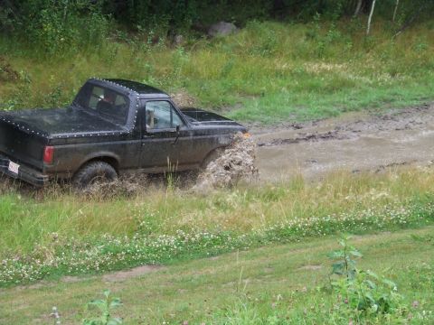 1990 Ford Bronco - A black 1990 Ford Bronco navigates a muddy trail, kicking up water and dirt.