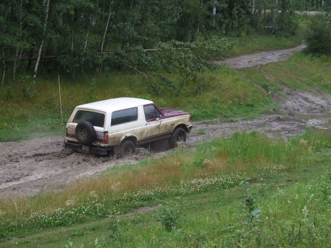 1990 Ford Bronco - A maroon and white Ford Bronco navigates a muddy track through a wooded area.