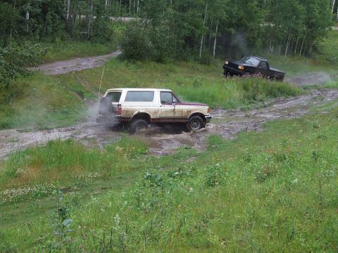 1990 Ford Bronco - A 1990 Ford Bronco navigates a muddy trail, kicking up water as a black pickup truck follows behind.