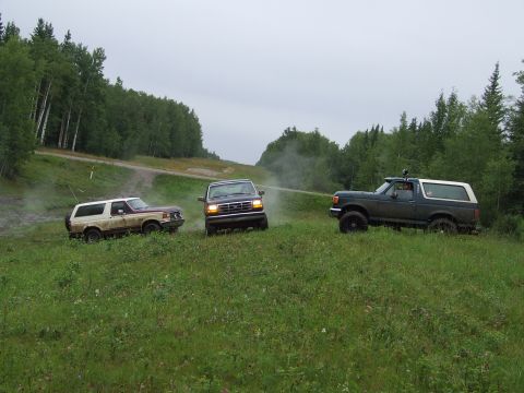 1990 Ford Bronco - Three Ford Broncos navigate a grassy, off-road terrain under an overcast sky.