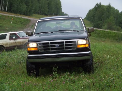 1990 Ford Bronco - Two Ford Broncos on a grassy, off-road path with trees in the background.