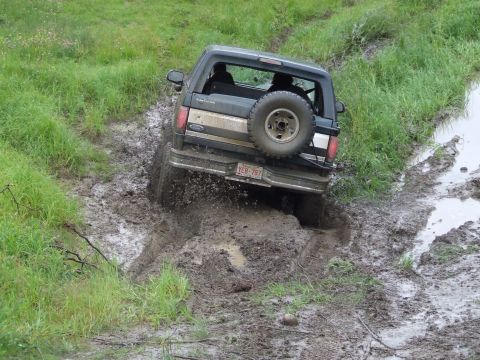 1990 Ford Bronco - A Ford Bronco navigates a muddy trail, kicking up dirt as it struggles for traction.