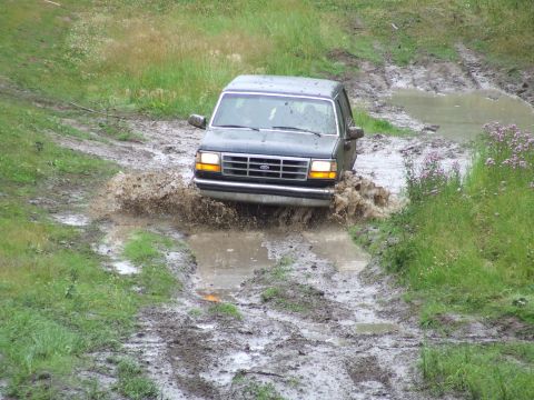 1990 Ford Bronco - A dark 1990 Ford Bronco navigates a muddy trail, kicking up water as it drives through a deep puddle.