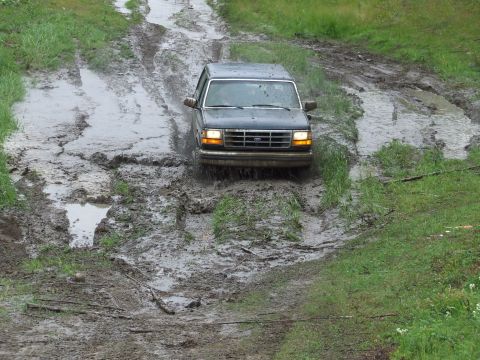 1990 Ford Bronco - A dark SUV navigates a treacherous muddy path, its front end angled downwards.