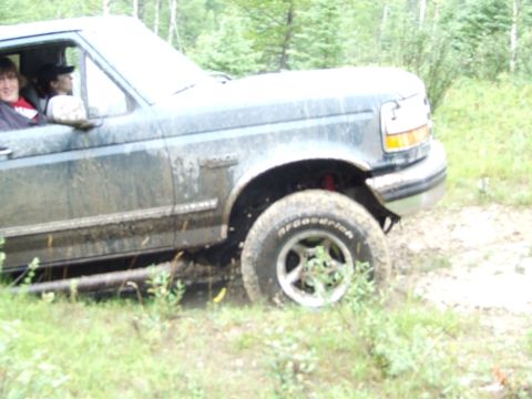 1990 Ford Bronco - A dirty Ford F-Series truck navigates a muddy, wooded trail with passengers visible inside the cab.