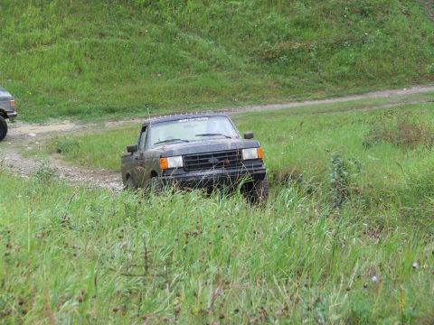 1990 Ford Bronco - A dark gray Ford F-Series truck navigates a grassy, off-road terrain with another vehicle partially visible to the left.