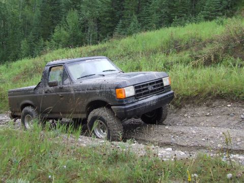 1990 Ford Bronco - A black 1990 Ford Bronco navigates a muddy, off-road trail surrounded by greenery.