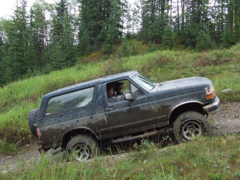 1990 Ford Bronco - A dark green 1990 Ford Bronco navigates a muddy off-road trail surrounded by trees.