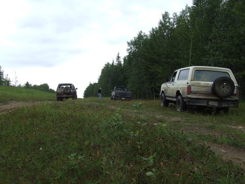 1990 Ford Bronco - Three off-road vehicles are parked on a dirt path surrounded by trees and grassy fields.