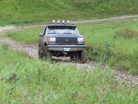 1990 Ford Bronco - A dark green 1987 Ford Bronco navigates a grassy, off-road trail with four lights mounted on its roof rack.