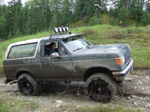 1990 Ford Bronco - A woman drives a dirty Ford Bronco with off-road tires and roof lights through a wooded area.