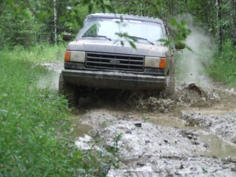 1990 Ford Bronco - A gray 1990 Ford Bronco navigates a muddy trail, kicking up water and dirt.
