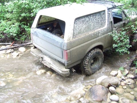 1990 Ford Bronco - A muddy Ford Bronco navigates a shallow, rocky stream surrounded by dense foliage.