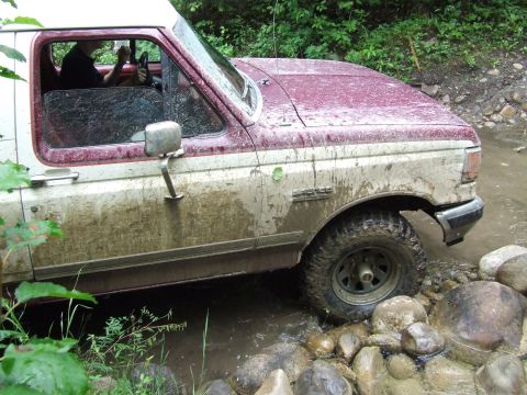 1990 Ford Bronco - Muddy 1990 Ford Bronco navigates a water crossing with large rocks.
