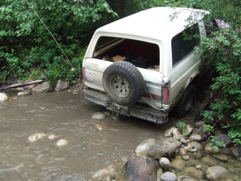 1990 Ford Bronco - A white Ford Bronco is partially submerged in a rocky stream, with trees and foliage surrounding it.