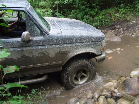 1990 Ford Bronco - A mud-splattered 1990 Ford Bronco navigates a rocky stream in a wooded area.