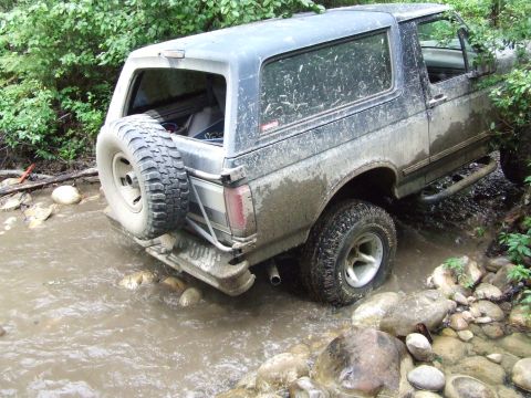 1990 Ford Bronco - Ford Bronco navigating a rocky stream bed surrounded by dense foliage.