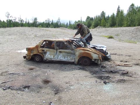 1990 Ford Bronco - A heavily damaged sedan, spray-painted with 'Burn Baby!', sits in a desolate area next to another vehicle.