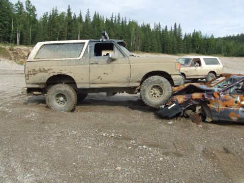 1990 Ford Bronco - A muddy 1990 Ford Bronco is parked on a gravel lot, with another SUV visible in the background.