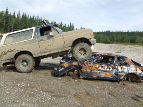 1990 Ford Bronco - A lifted Ford Bronco is parked on top of a heavily damaged and rusted car in a muddy outdoor area.