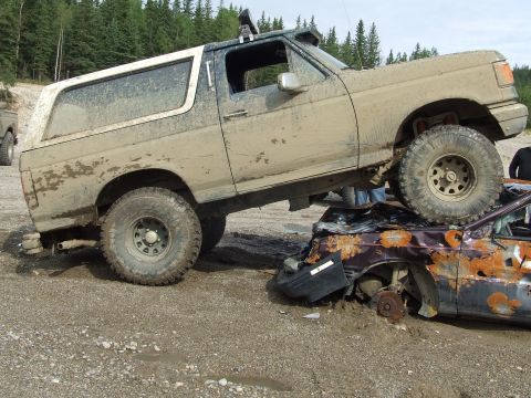1990 Ford Bronco - A muddy Ford Bronco is driven over a heavily damaged car in a wooded area.