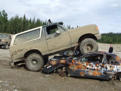 1990 Ford Bronco - A lifted Ford Bronco is driven over a heavily damaged car, with another vehicle visible in the distance.