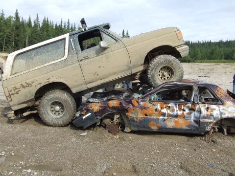 1990 Ford Bronco - A Ford Bronco is perched atop a heavily damaged car, both covered in mud and debris.