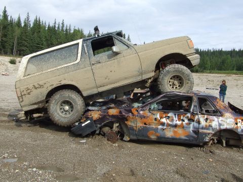 1990 Ford Bronco - 1990 Ford Bronco crushing a wrecked car in a dirt lot with trees in the background.