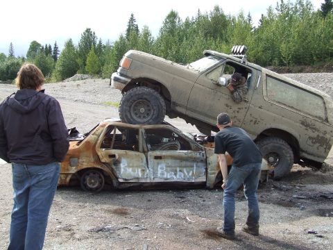 1990 Ford Bronco - A modified Ford Bronco is parked on top of a heavily damaged and burnt sedan, with two onlookers observing the scene.