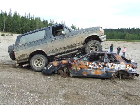 1990 Ford Bronco - A muddy 1990 Ford Bronco is parked on top of a heavily damaged car in a rural outdoor setting.
