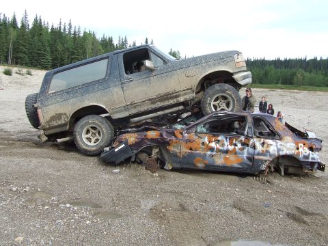 1990 Ford Bronco - A 1990 Ford Bronco is parked on top of a destroyed car in a dirt lot.