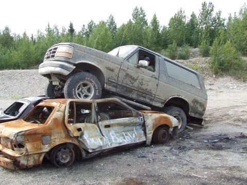 1990 Ford Bronco - A 1992 Ford Bronco is parked on top of a severely damaged and rusted sedan in a junkyard.