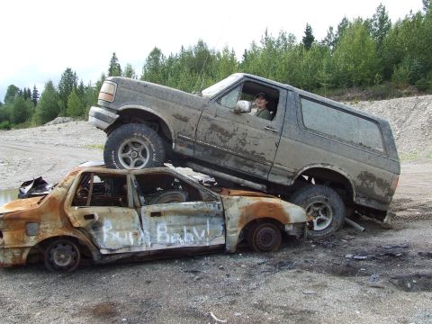 1990 Ford Bronco - A 1990 Ford Bronco is parked on top of a burnt-out sedan that has "Burn Baby!" spray-painted on its side.