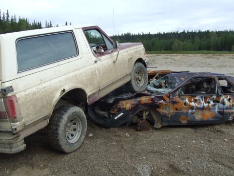 1990 Ford Bronco - A white and maroon Ford Bronco is parked on top of a heavily damaged and rusted car in a junkyard.