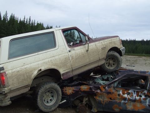 1990 Ford Bronco - A 1990 Ford Bronco is piled on top of a heavily damaged car in a junkyard.