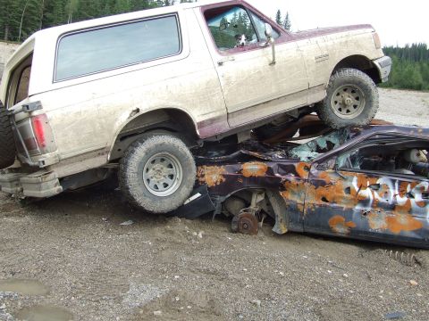 1990 Ford Bronco - A Ford Bronco is perched atop a heavily damaged and rusted car, with a dog looking out from the Bronco's window.