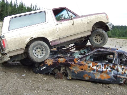 1990 Ford Bronco - A 1990 Ford Bronco rests atop a heavily damaged car in a junkyard setting.