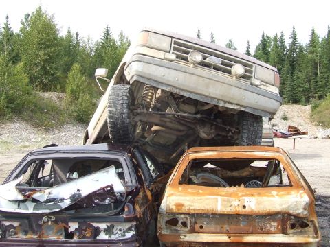 1990 Ford Bronco - A tan Ford truck is precariously perched atop two heavily damaged cars in a junkyard.